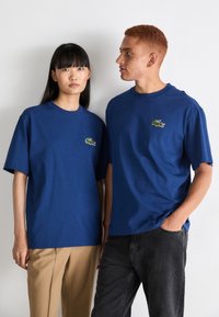 Camisetas de algodón azul con manga corta, que presentan un logo de cocodrilo verde bordado en el lado izquierdo del pecho, llevadas con pantalones beige y negros.
