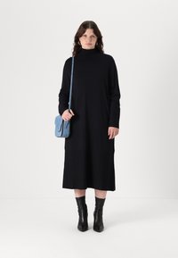 Abito nero lungo con maniche lunghe e colletto alto, abbinato a una borsa a tracolla blu. Stivaletti neri completano il look. Sfondo neutro.