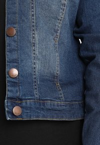 Denim-takki haalistuneessa sinisessä värissä, varustettuna metallinapeilla, tummalla ompeleella ja klassisella kaulustyylillä, tuoden esiin kankaan tekstuuritiedot.