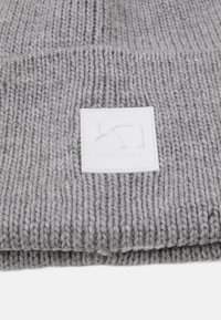 Grå stickad mössa med tjock textur, prydd med en vit fyrkantig lapp med ett präglat logotyp. Enkla, flexibla design som passar för kallt väder.