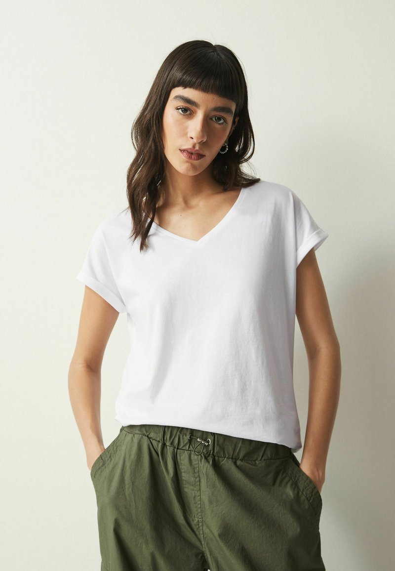 T-shirt bianca in cotone con scollo a V e maniche corte arrotolate, abbinata a pantaloni leggeri con vita elastica di colore verde oliva.