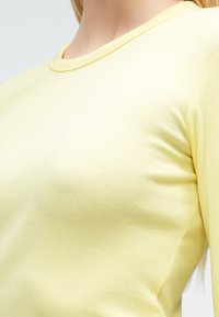 Gul ribbestrikket langermet genser med rund hals. Teksturen er glatt med subtile vertikale striper som fremhever en tettsittende design.