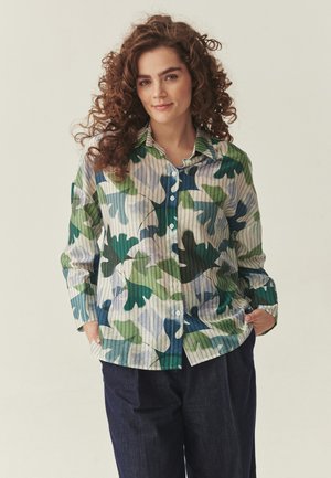 Mlada žena s kovrčavom smeđom kosom, odjevena u zelenu, plavu i bež košulju s uzorkom na kopčanje i tamne hlače, ruke u džepovima, blago se smiješi.