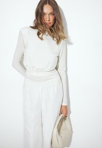 Weißes Langarmoberteil mit geraffter Taille, kombiniert mit flie�ßenden weißen Hosen. Das Model hält eine strukturierte hellfarbene Tasche.