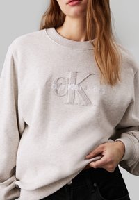 Sudadera de algodón gris claro con escote redondo, puños acanalados y logo bordado de "Calvin Klein Jeans" en textura suave.