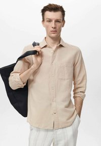 Light beige linen shirt with a chest pocket, button front, and rolled sleeves. Model holds a black tote bag. White striped pants.