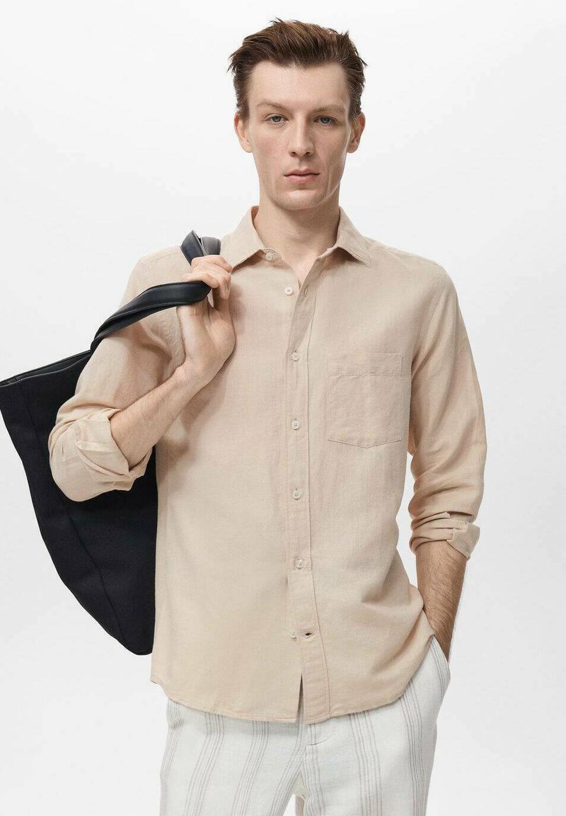 Light beige linen shirt with a chest pocket, button front, and rolled sleeves. Model holds a black tote bag. White striped pants.