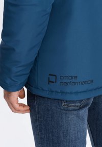 Modrá zateplená bunda s matným povrchem, se znakem v černé barvě blízko lemu. Spárovaná s modrými džínami.