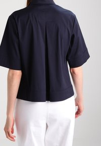 Navyblå kortärmad skjorta med krage, med en detalj av en bakveck och rakt fåll. Tillverkad av mjukt tyg, i kombination med vita byxor.