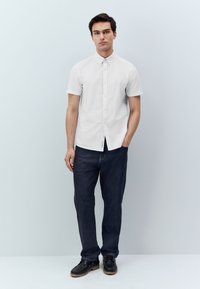 Chemise blanche à manches courtes avec boutons, associée à un jean foncé et des chaussures noires. Le modèle pose avec une main dans la poche, sur un fond clair.