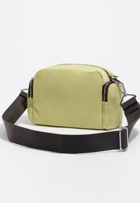 Rectangular, light green bag made of soft material, featuring a zipped top, brown adjustable strap, and silver hardware accents.