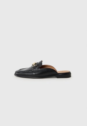 Mocasín negro de cuero sin talón, con detalle de hebilla dorada y tacón plano bajo sobre un fondo neutro.