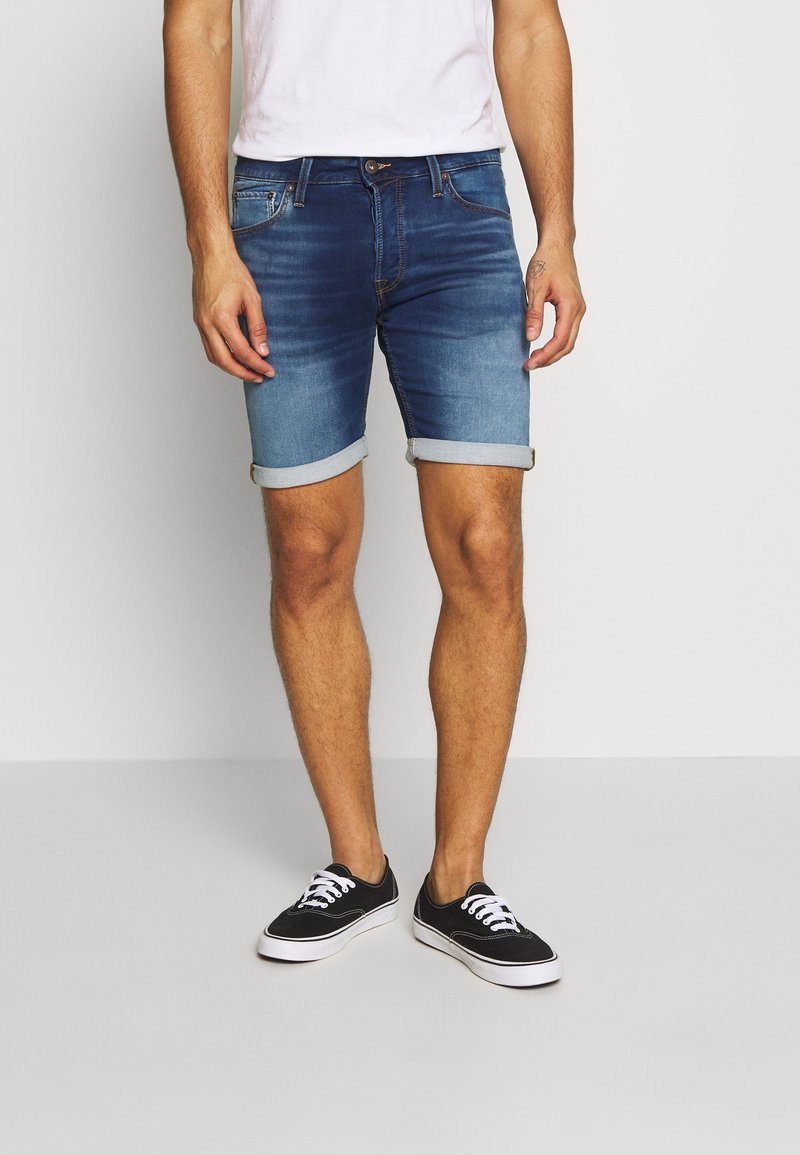 Shorts de denim en un lavado azul medio con un aspecto ligeramente desgastado, dobladillos enrollados y diseño estándar de cinco bolsillos, combinados con zapatillas negras.