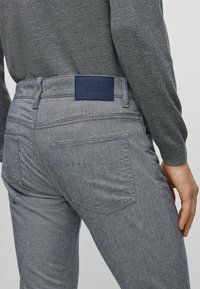 Jean en denim gris avec une finition texturée, présentant une patte de cuir bleu foncé frappée du logo "BOSS", et des poches arrière standard.
