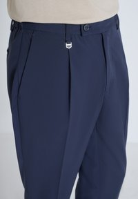 Marineblauwe broek met voorplooien, zachte textuur en een zilveren metalen sluiting bij de taille. Knopsluiting op de tailleband.