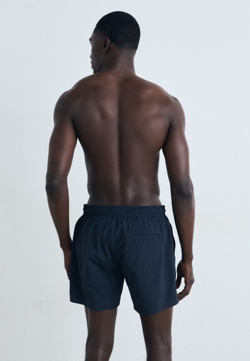 Marinefarbene Schwimmshorts mit elastischem Bund und einer Gesäßtasche, von hinten gezeigt, das Modell hat einen muskulösen Körperbau und kurz geschnittenes Haar.