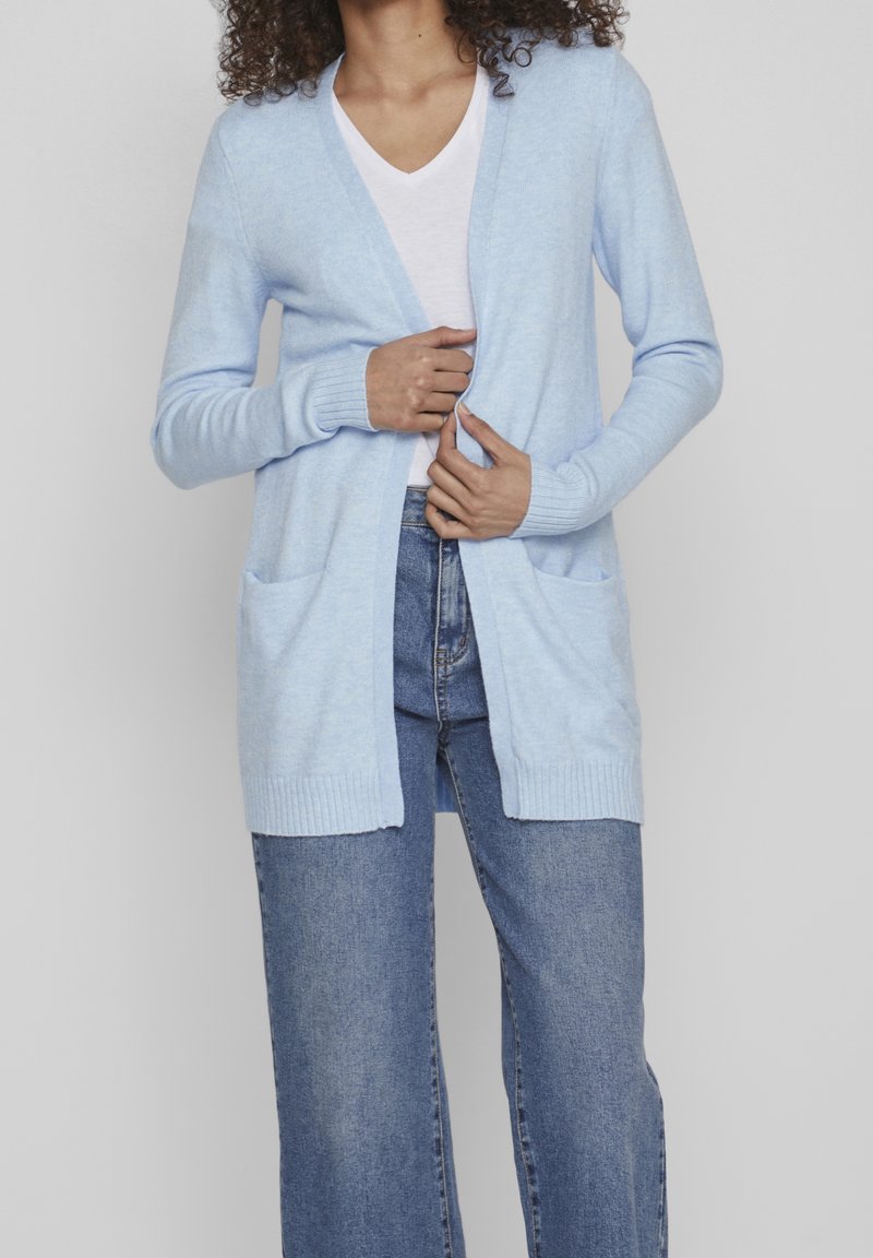 Femme portant un cardigan bleu clair ouvert devant sur un t-shirt blanc col en V et un jean bleu taille haute, ajustant l'avant du cardigan.