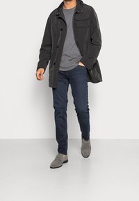 Mörkgrå lättviktsjacka med en framficka, tillsammans med en grå t-shirt, mörka jeans och grå loafers. Modell som går på en jämn yta.