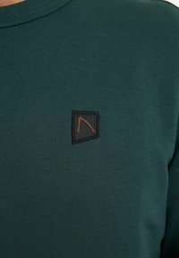 Felpa verde scuro con lavorazione a maglia strutturata, scollatura rotonda e una piccola toppa nera con dettaglio logo arancione.
