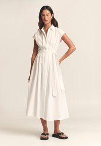 Witte, knielange overhemdjurk met korte mouwen, tailleband en een soepel vallende rok. Gecombineerd met zwarte platte sandalen. Soepele stoftextuur.