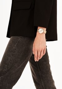 Montre-bracelet avec un bracelet en maille dorée et un cadran argenté, portée au poignet. Un blazer noir texturé et un jean gris foncé sont également visibles.