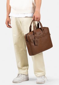 Bruine leren aktetas met dubbele handgrepen, gestructureerd oppervlak, logo-detail en een label. Draag je metlichte crèmekleurige broeken en witte sneakers.