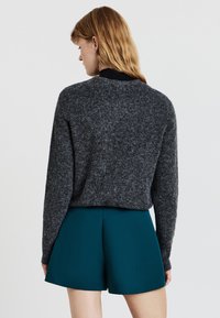 Donna con i capelli castano chiaro, che indossa un maglione grigio scuro e pantaloni corti teal a vita alta, vista da dietro su uno sfondo semplice.