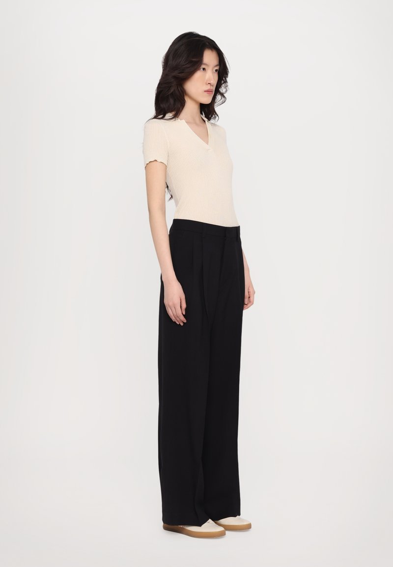 Ribbed, cream short-sleeve top with V-neck, paired with high-waisted, black wide-leg pants. The model stands sideways, wearing beige shoes.