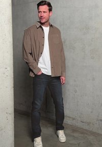 Mann steht drinnen, trägt ein braunes Hemd mit Knöpfen, ein weißes T-Shirt, dunkle Jeans und weiße Sneakers vor einer grauen Betonwand.
