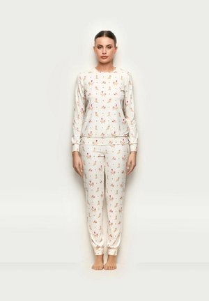 Mujer descalza, con camiseta de manga larga y pantalones blancos con pequeños patrones de perros y flores sobre un fondo liso.