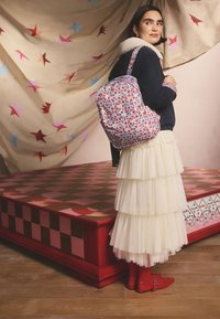 Femeie cu păr scurt și închis la culoare purtând o jachetă bleumarin, o fustă crem în straturi, pantofi roșii și un rucsac cu model floral, stând lângă o platformă cu model în carouri roșii.