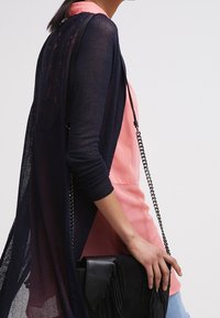 Marinblå mesh-kardigan över en rosa topp, i kombination med en svart fransad handväska och ljusblå jeans, som har en kedjeslinger och texturerad tyg.