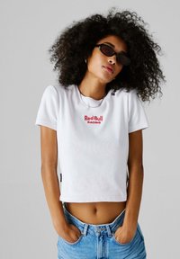 Νεαρό άτομο με σγουρά μαλλιά, φοράει μαύρα γυαλιά ηλίου, λευκό κοντό μπλουζάκι "Red Bull Racing" και μπλε τζιν με τα χέρια στις τσέπες.