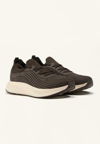 Een paar donkerbruine gebreide sneakers met golvenpatronen en dikke witte zolen, schuin naar rechts op een effen lichte achtergrond.