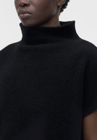 Gros plan d'une personne portant un pull noir texturé à col montant avec des manches courtes sur un fond uni.