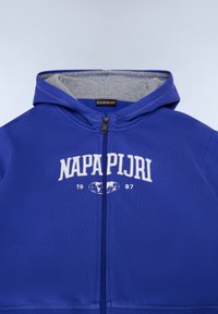 Sudadera con cremallera azul con interior gris, que presenta un logo blanco y el año '1987' en el pecho en una fuente audaz. Textura de tela suave.