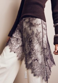Osoba oblečená v bielych nohaviciach s čiernou kvetinovou čipkovou vrstvou a tmavohnedom rebrovanom svetri, s náramkami na rukách a uvoľneným postojom.