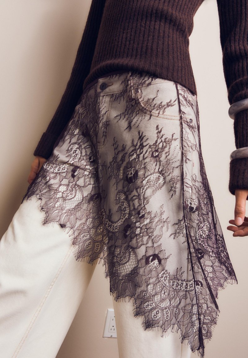 Osoba oblečená v bielych nohaviciach s čiernou kvetinovou čipkovou vrstvou a tmavohnedom rebrovanom svetri, s náramkami na rukách a uvoľneným postojom.