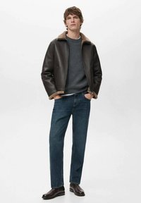 Giubbotto in pelle marrone con colletto in shearling beige, indossato sopra un maglione grigio, abbinato a jeans blu e stivali marrone scuro. Design dritto.