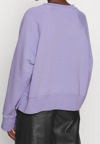 Sudadera de un ligero tono púrpura con un corte holgado, cuello acanalado y mangas raglán. Presenta un dobladillo redondeado y una textura sutil.