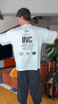 Άνδρας στέκεται μέσα σε κλειστό χώρο με τεντωμένα χέρια, φοράει ένα λευκό μπλουζάκι με μαύρο κείμενο και σύμβολα στην πλάτη και φαρδιά σκούρα παντελόνια.