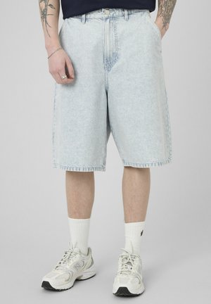 Homme portant un short en denim bleu clair, des baskets blanches et des chaussettes blanches montantes, debout les mains dans les poches devant un fond uni.