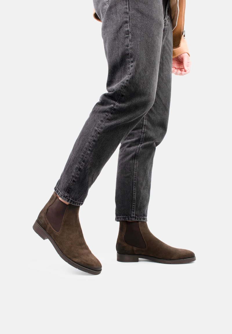 Botas Chelsea de ante marrón con paneles laterales elásticos, puntera redondeada y tacón bajo apilado, combinadas con jeans gris oscuro.