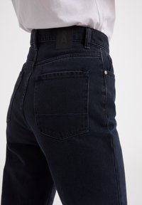 Jean en denim bleu foncé avec une coupe taille mi-haute, des poches arrière et une étiquette noire rectangulaire sur la ceinture. Texture lisse et design élégant.