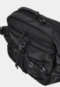 Sac en tissu noir avec plusieurs compartiments zippés, des sangles réglables et des accents en cordon élastique, arborant un logo sur le devant.