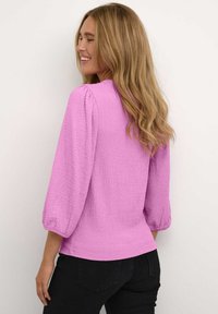 Blusa textured viola chiaro con maniche a tre quarti, scollo rotondo e orlo dritto, abbinata a pantaloni neri per un contrasto.