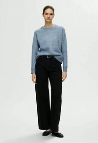 Mulher de pé, vestindo um suéter azul claro, calças largas pretas com cinto e sapatos planos pretos, contra um fundo simples.