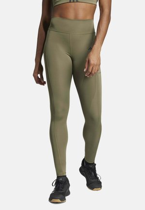 Vrouw draagt olijfgroene leggings met hoge taille en zwarte sportschoenen, staand voor een effen achtergrond.