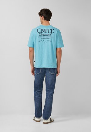 Man gezien van achteren, draagt een lichtblauw T-shirt met de tekst "UNITE Connect", donkere blauwe jeans en witte sneakers tegen een effen grijze achtergrond.