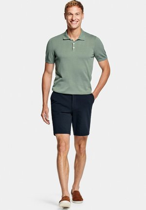 Smiling man walking, wearing light green short-sleeve polo shirt, black shorts, and brown slip-on shoes against a plain white background.